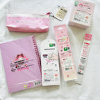 A flat lay of Sanrio My Melody stationery items on a white fabric background, including a pink zip pouch, a pink spiral notebook, a white pen case, and two packaged pens.