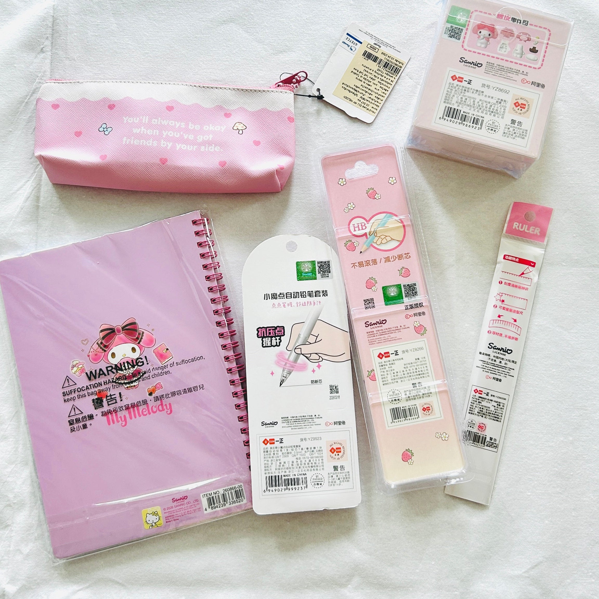 A flat lay of Sanrio My Melody stationery items on a white fabric background, including a pink zip pouch, a pink spiral notebook, a white pen case, and two packaged pens.