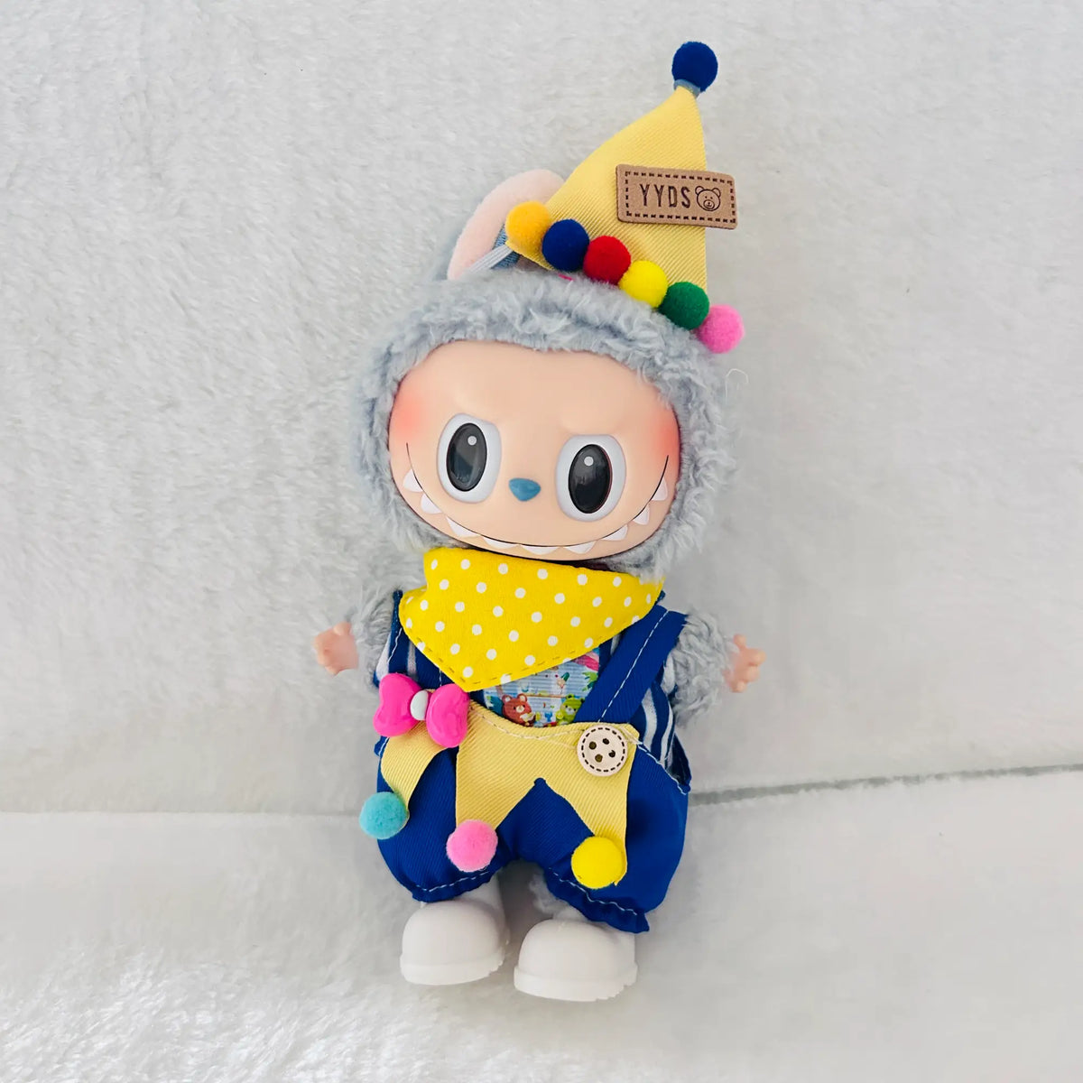 A labubu plush doll has a yellow party hat with colorful pom-poms, a yellow polka-dot bandana, and blue overalls.