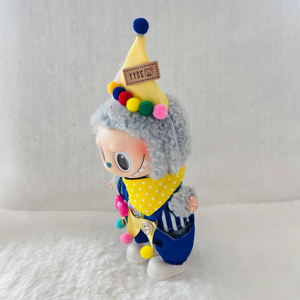 A Labubu doll wearing a yellow pointed hat with colourful pom-poms, a yellow polka-dot bandana, and blue striped pants.