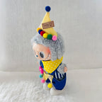 A Labubu doll wearing a yellow pointed hat with colourful pom-poms, a yellow polka-dot bandana, and blue striped pants.