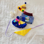 A flat-lay of a Labubu doll's costume set includes a blue and white striped top, a colourful pom-poms hat and overall, and a yellow bib.