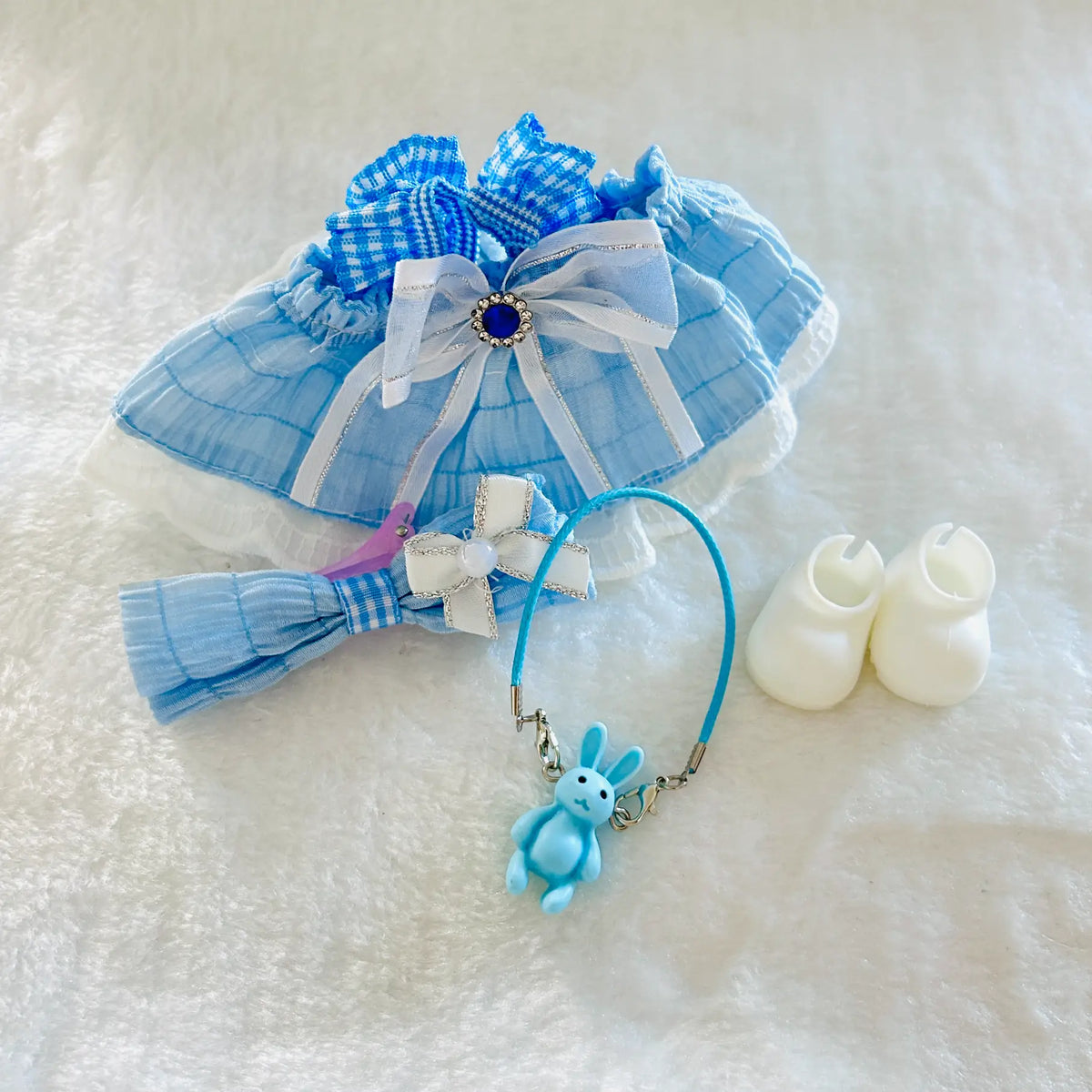 A front view of doll outfit set ncluding a light blue and white gingham ruffled dress with white lace trim, a matching headband with a pink bow, a blue keychain with a bunny charm.