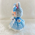 A small plush doll wearing a light blue dress with white lace trim, photographed against a soft white fabric background.