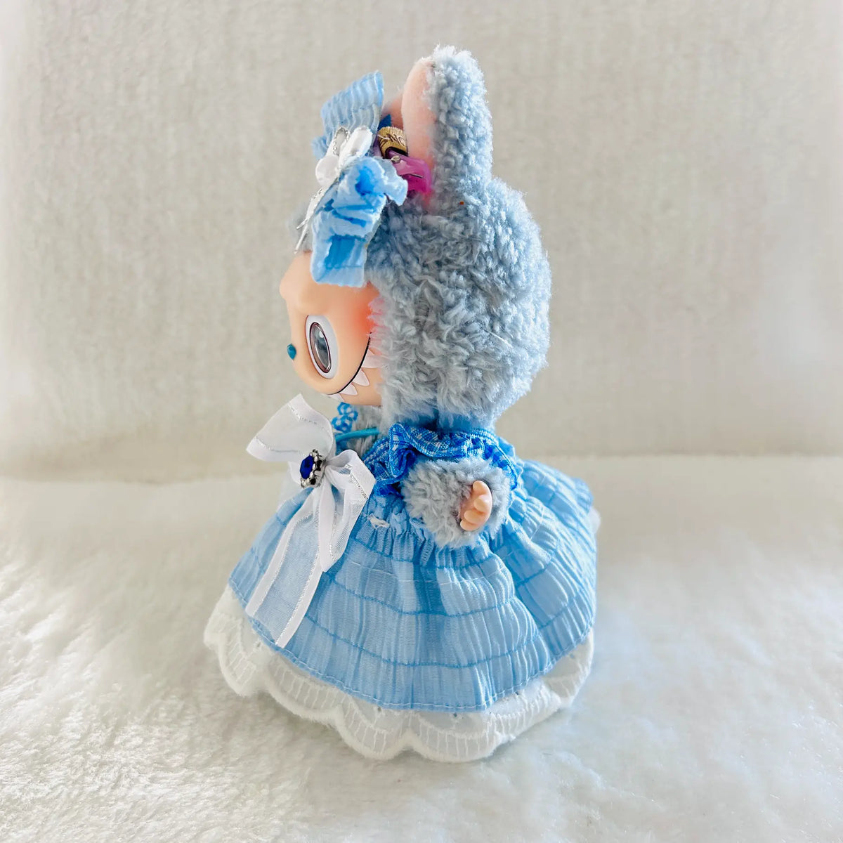 A small plush doll wearing a light blue dress with white lace trim, photographed against a soft white fabric background.
