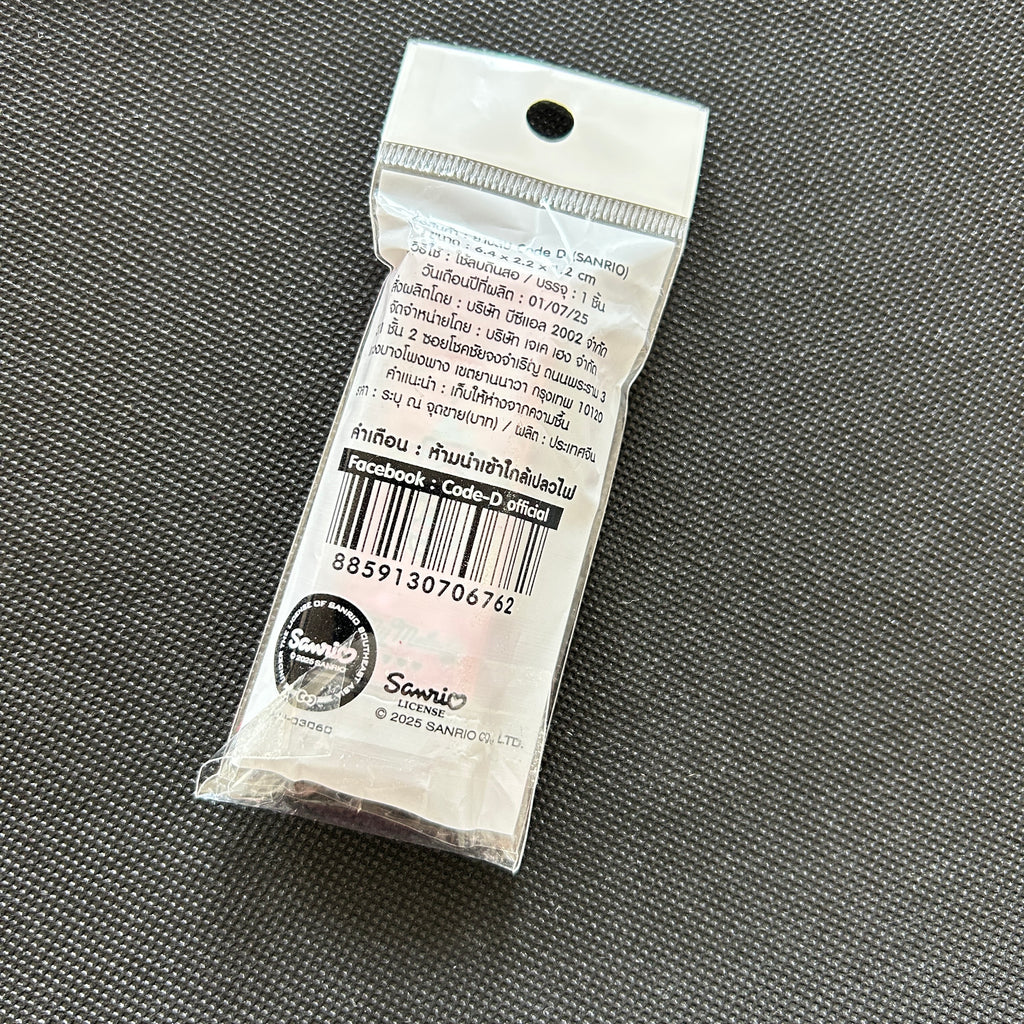 A small sealed white plastic package containing a single round coin or medallion, photographed on a dark textured fabric background. The package has printed text, a barcode, and a visible Sanrio logo near the bottom.