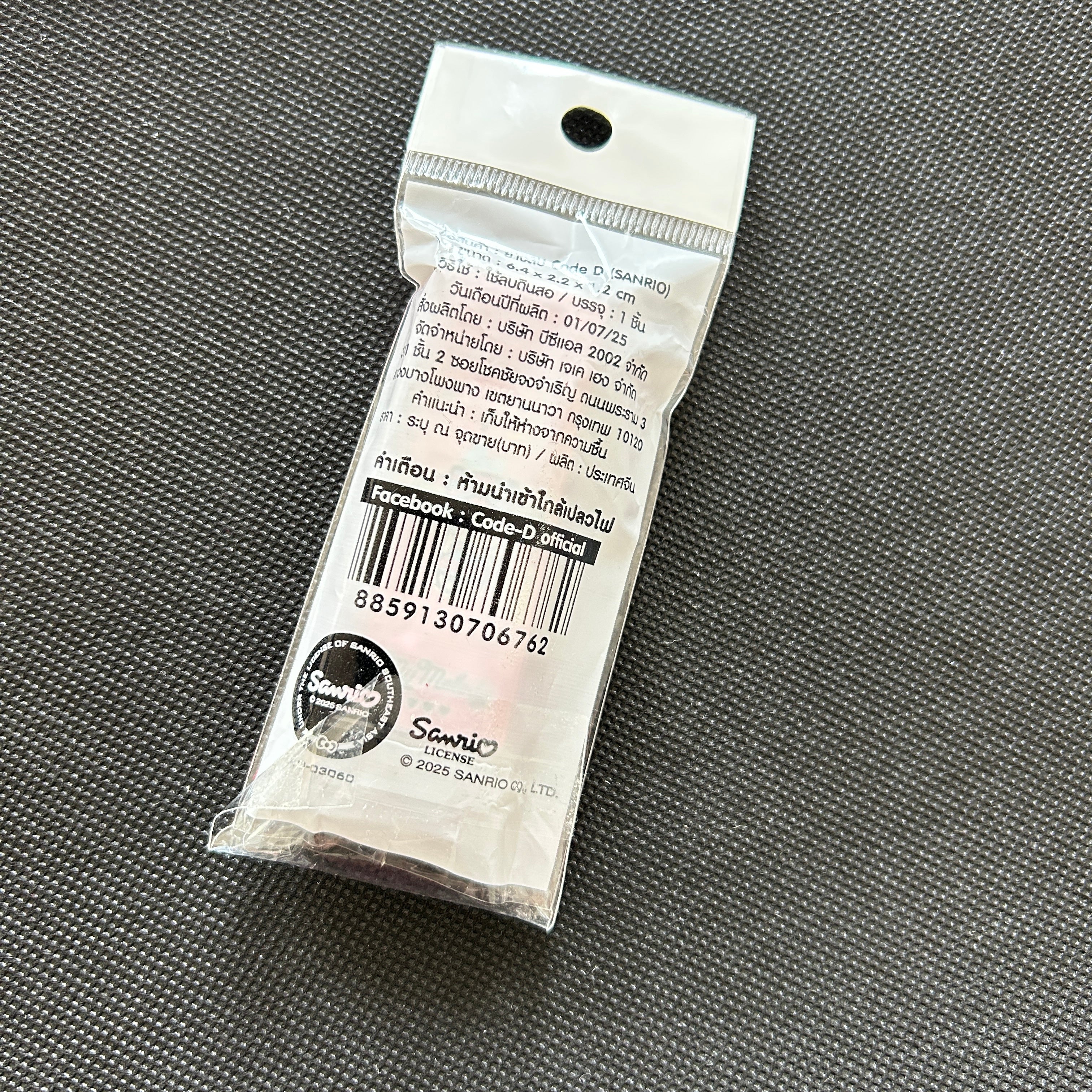 A small sealed white plastic package containing a single round coin or medallion, photographed on a dark textured fabric background. The package has printed text, a barcode, and a visible Sanrio logo near the bottom.
