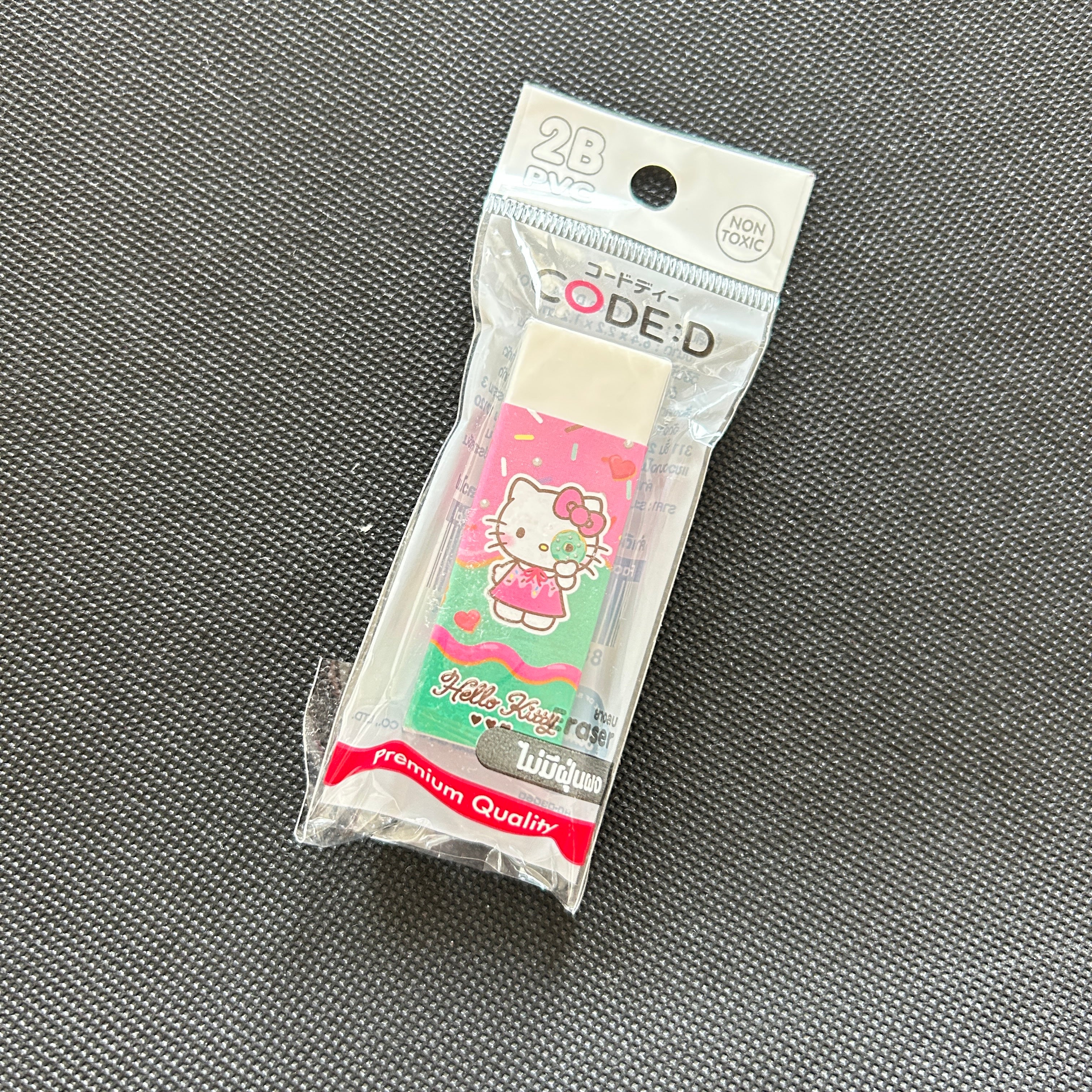 A packaged Hello Kitty eraser on a textured gray fabric background. The eraser is sealed in a clear plastic bag with a printed header card that reads 'CODE:D' and '28 TOYS', featuring a pink and green Hello Kitty design.