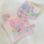 A set of pastel pink and purple Little Twin Stars stationery items, including a lined notepad, a washi tape roll, and several small cards or stickers, arranged on a soft white textured fabric background.