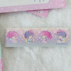 A close-up of a Sanrio character tape or sticker strip featuring two cute cartoon girls in pink and purple outfits, placed on a soft white faux-fur surface with pink star-patterned paper partially visible in the background.