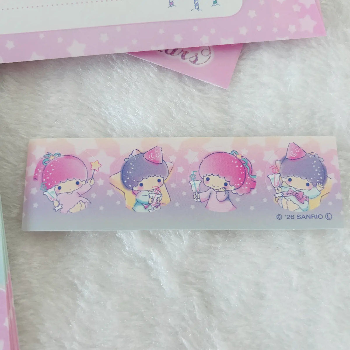 A close-up of a Sanrio character tape or sticker strip featuring two cute cartoon girls in pink and purple outfits, placed on a soft white faux-fur surface with pink star-patterned paper partially visible in the background.