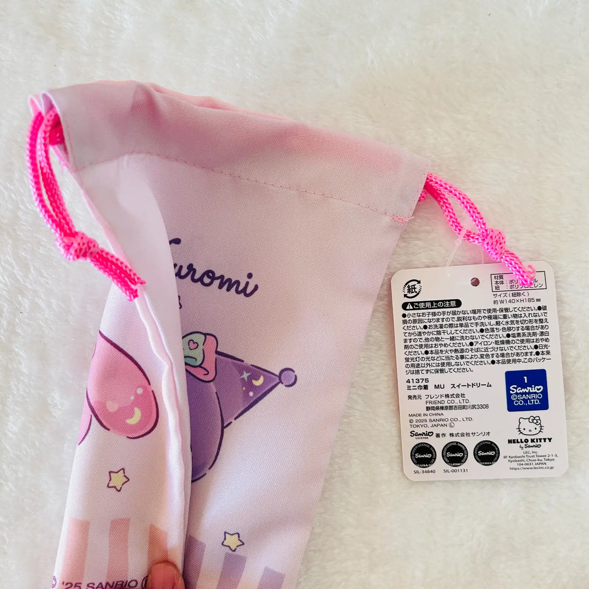 A pink drawstring pouch with a cartoon character print and a Sanrio hangtag, photographed on a soft white textured fabric background.