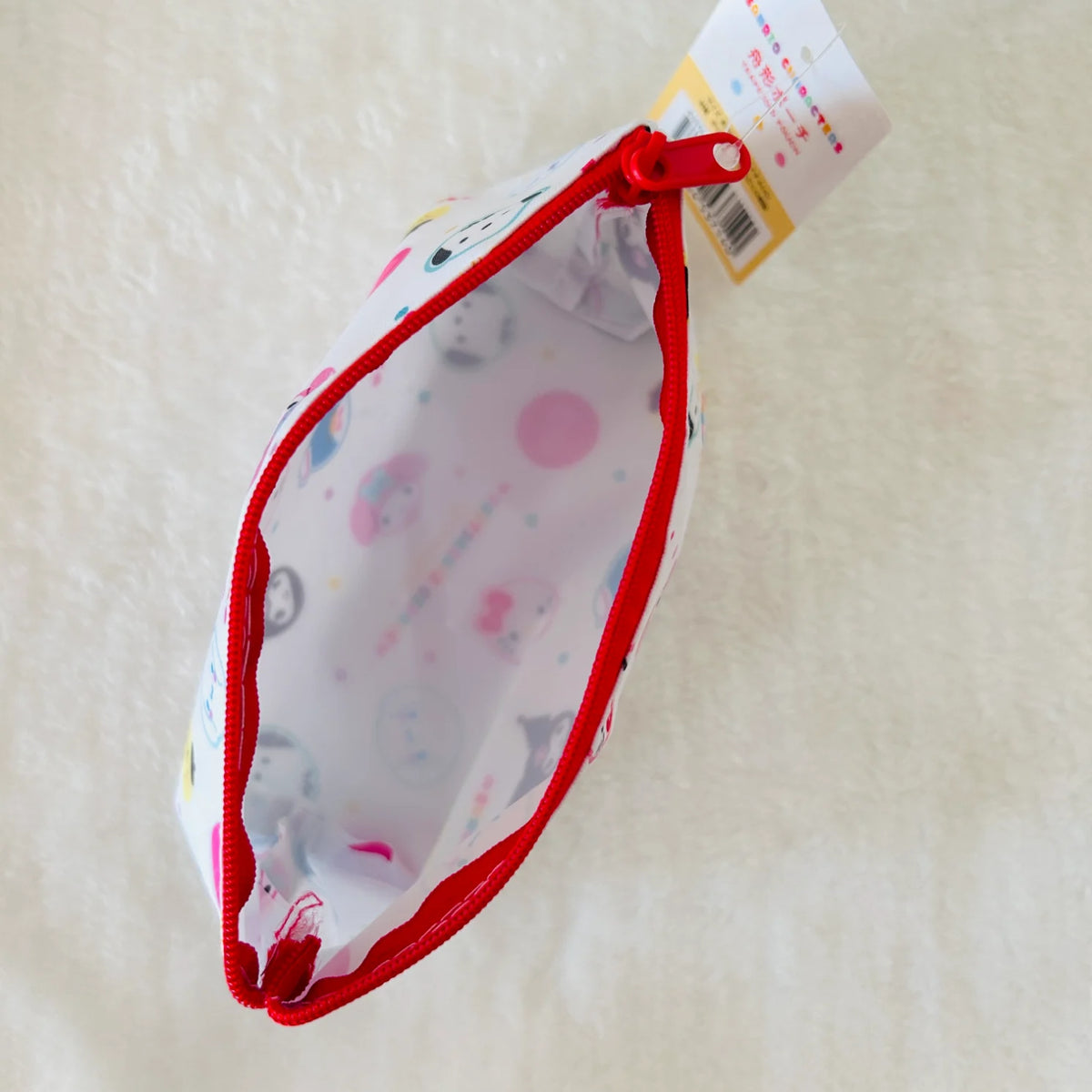 A small zippered pouch with a white background and a multicolor cartoon print, featuring a bright red zipper trim and a red pull tab. The pouch is shown open with its product tag still attached, set against a soft, textured off-white background.