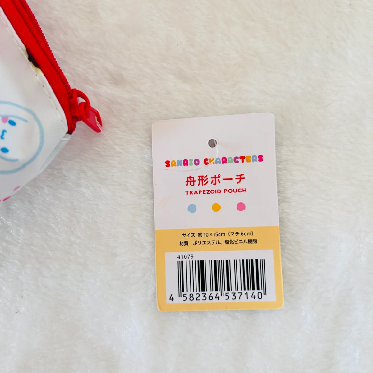 A product tag for a Sanrio Characters Trapezoid Pouch, placed on a white textured surface next to a partial view of a white and red zippered pouch.