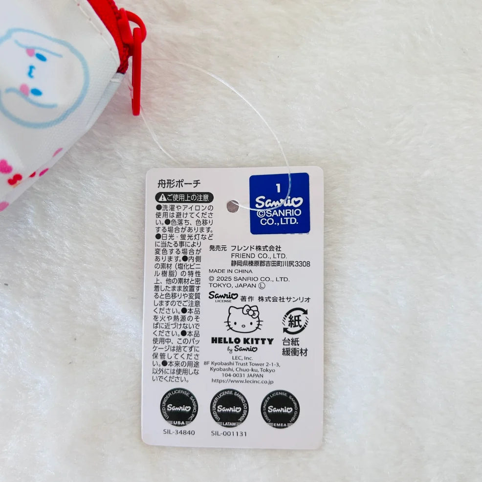 A close-up of a white product tag for a Sanrio Hello Kitty item, with Japanese text, the Sanrio logo, and Hello Kitty branding, placed on a white textured surface. Part of a white item with a red drawstring and blue cartoon face is visible at the top left.