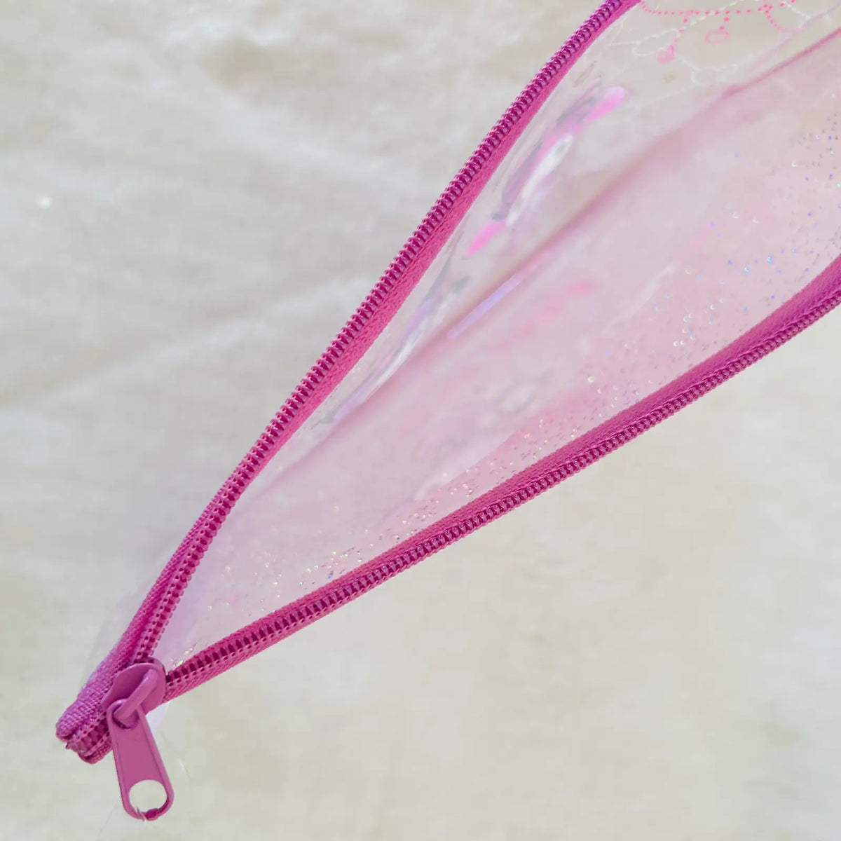 A close-up of a clear pencil pouch with pink glitter and a bright pink zipper, shown on a light beige textured background.