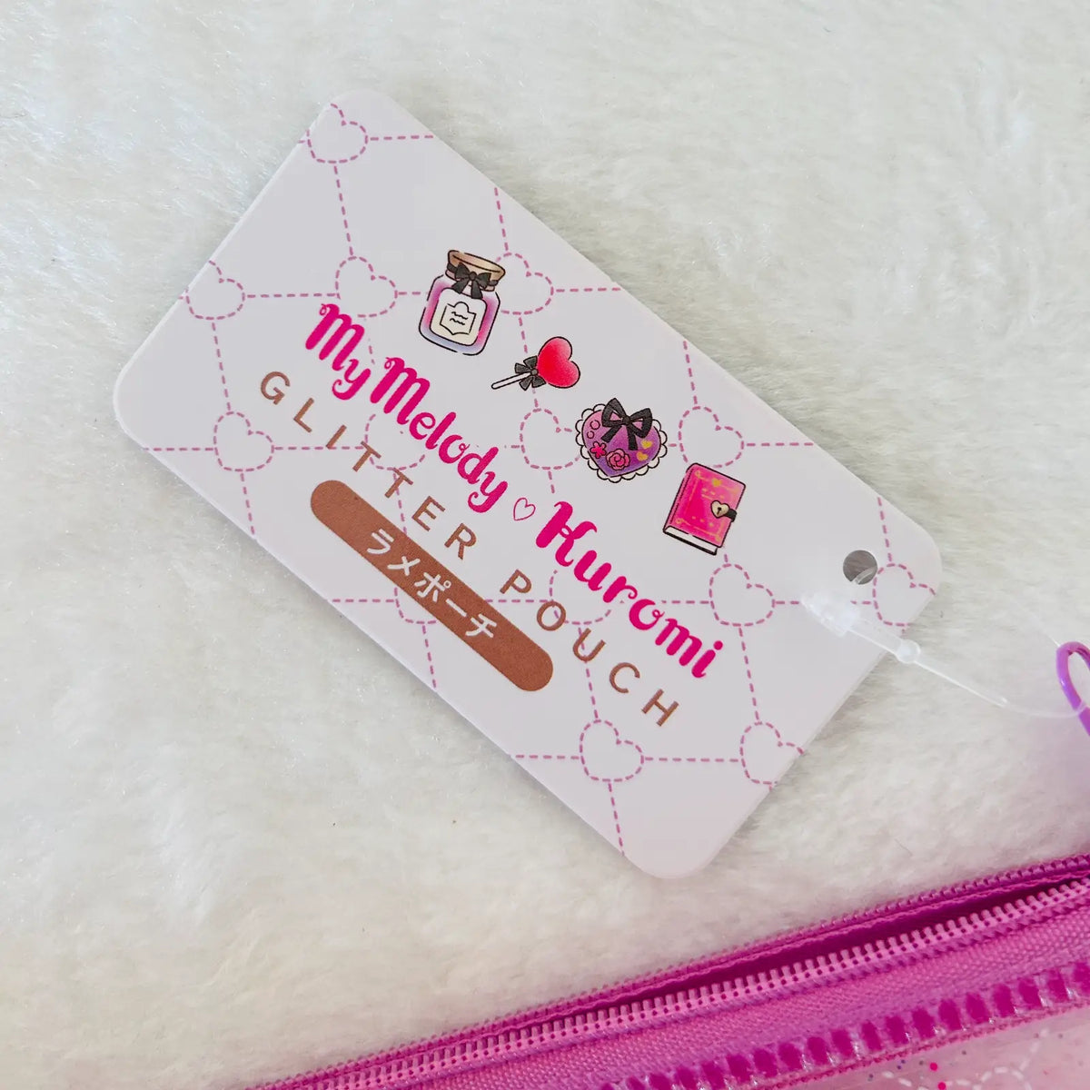 A close-up of a pink glitter pouch with its attached paper tag. The tag is white with pink dashed heart outlines, featuring the text 'My Melody Huromi GLITTER POUCH' and Japanese characters. 