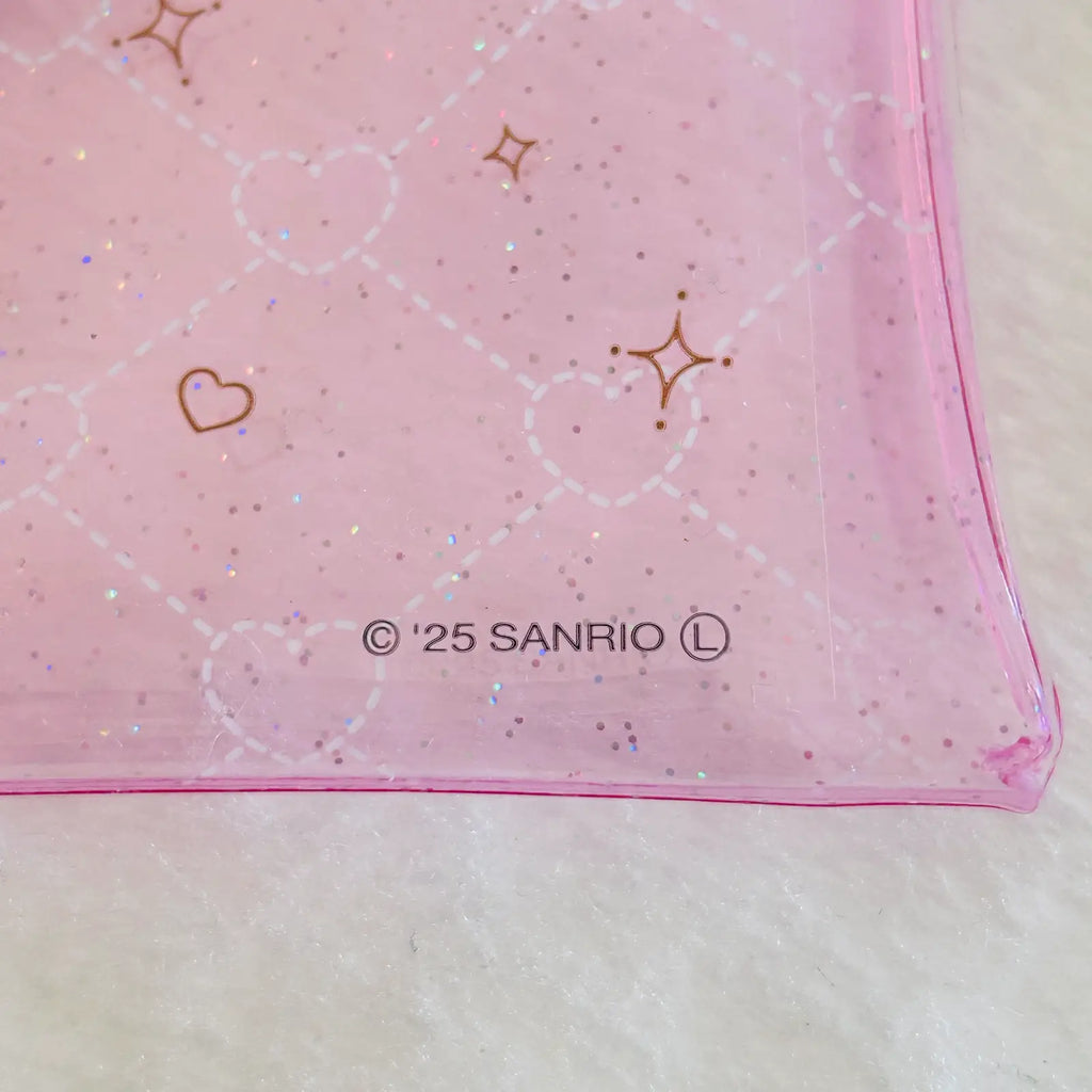 A close-up of a pink translucent plastic pouch with a white heart lattice pattern, gold heart and star outlines, and '© 25 SANRIO' printed near the bottom, resting on a white textured surface.
