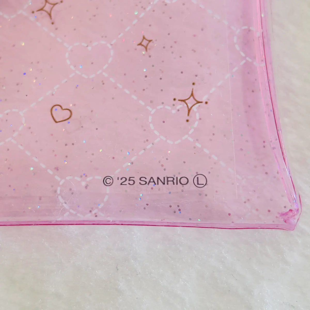 A close-up of a pink translucent plastic pouch with a white heart lattice pattern, gold heart and star outlines, and '© 25 SANRIO' printed near the bottom, resting on a white textured surface.