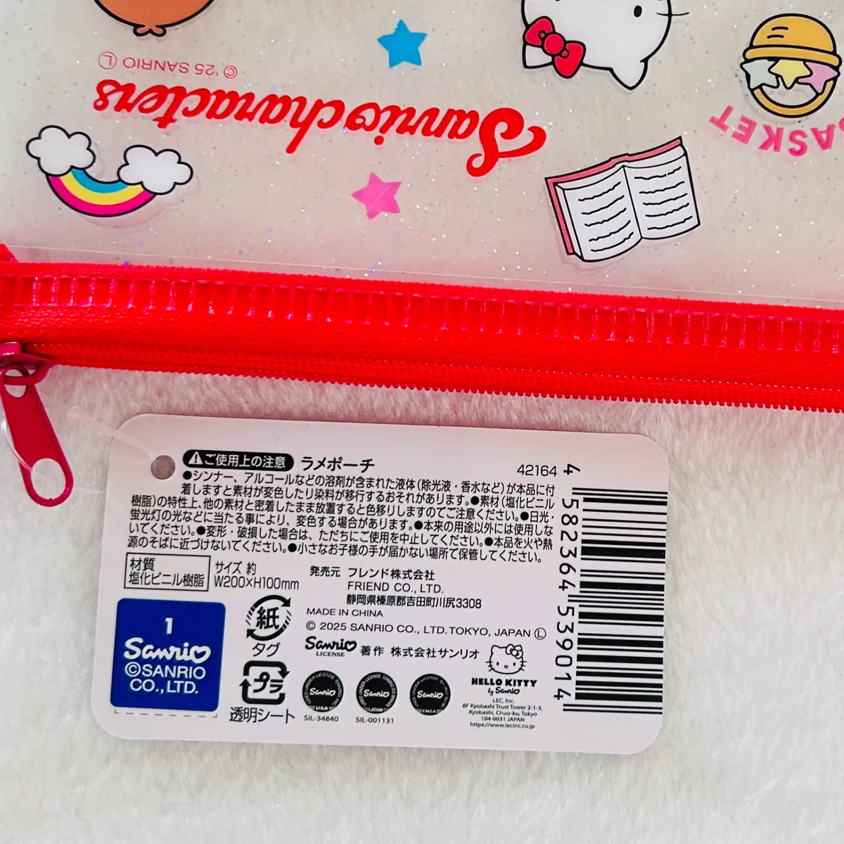 A close-up of a white glittery Sanrio Hello Kitty zippered pouch with a bright pink zipper, featuring printed Sanrio and Hello Kitty graphics, alongside a white product tag with Japanese text and a barcode.