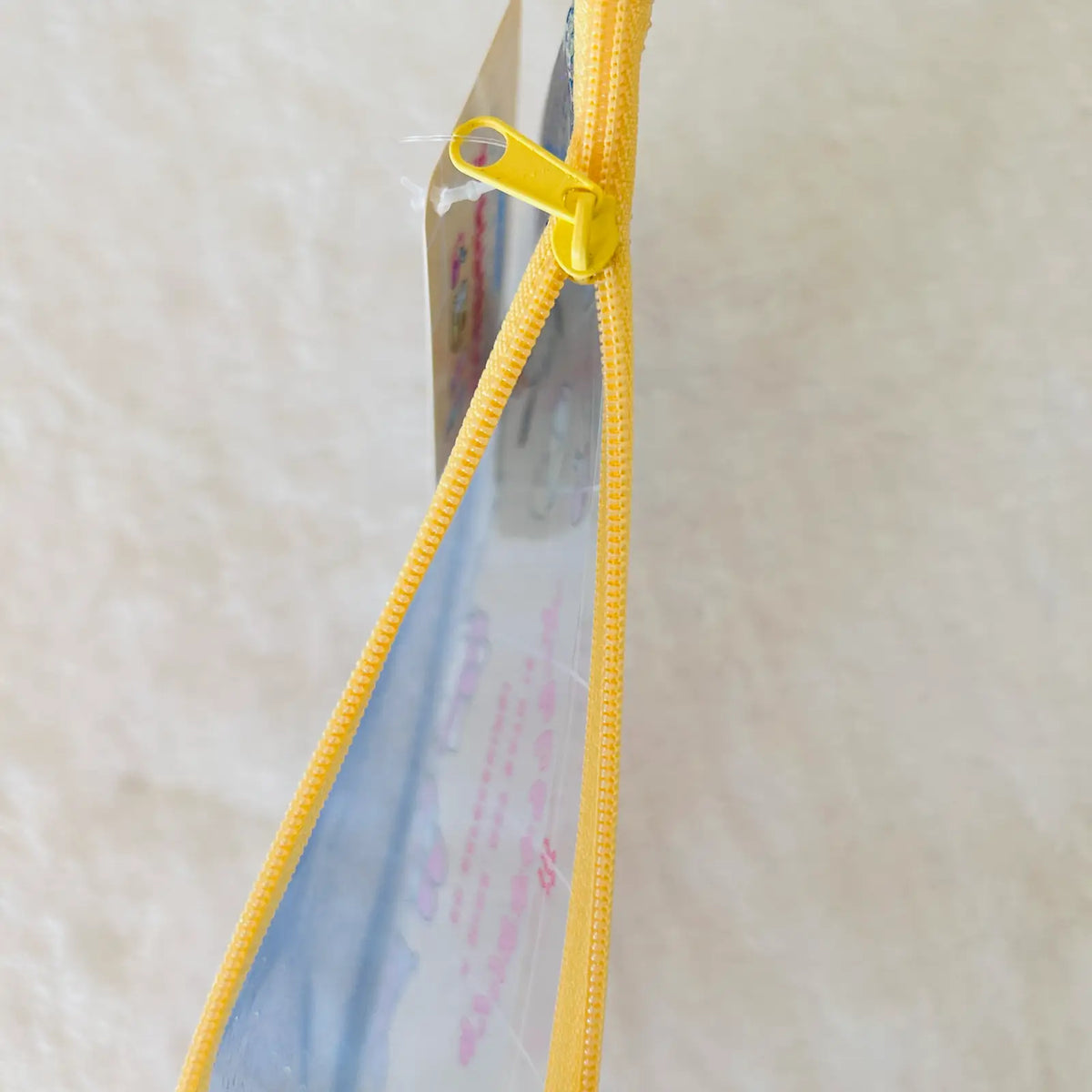 A close-up of a clear plastic zipper pouch with a bright yellow zipper and yellow pull tab, shown against a light beige textured background.