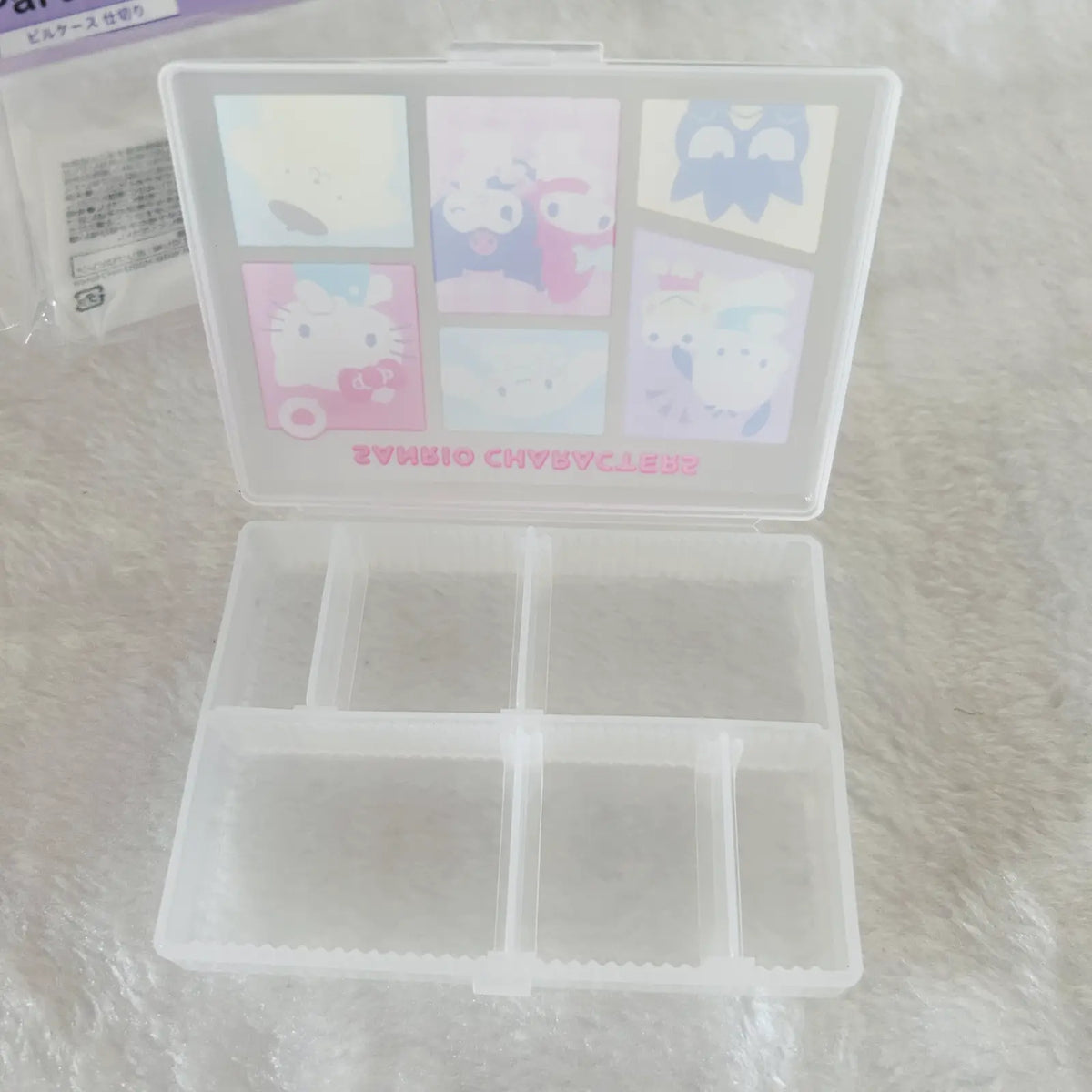 A translucent white plastic organizer box with six compartments in the base and a hinged lid decorated with pastel Sanrio character illustrations, including Hello Kitty, shown on a white textured surface.