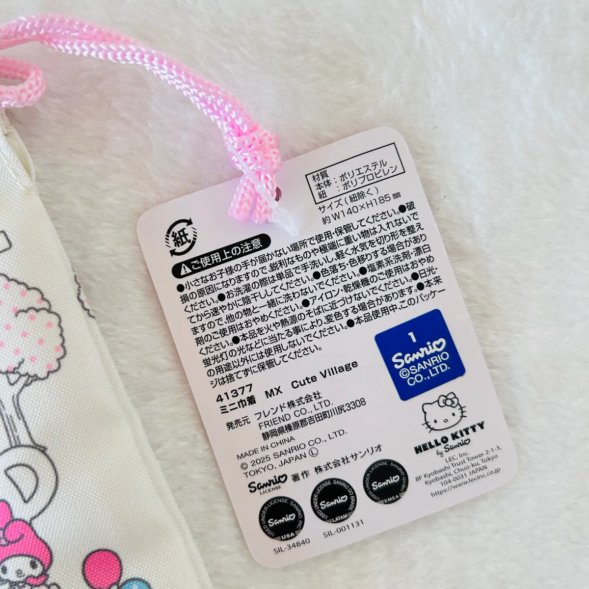 A close-up of a Hello Kitty product tag attached with a pink braided cord, showing Japanese text, product code 41577, and Sanrio branding on a white fabric background.