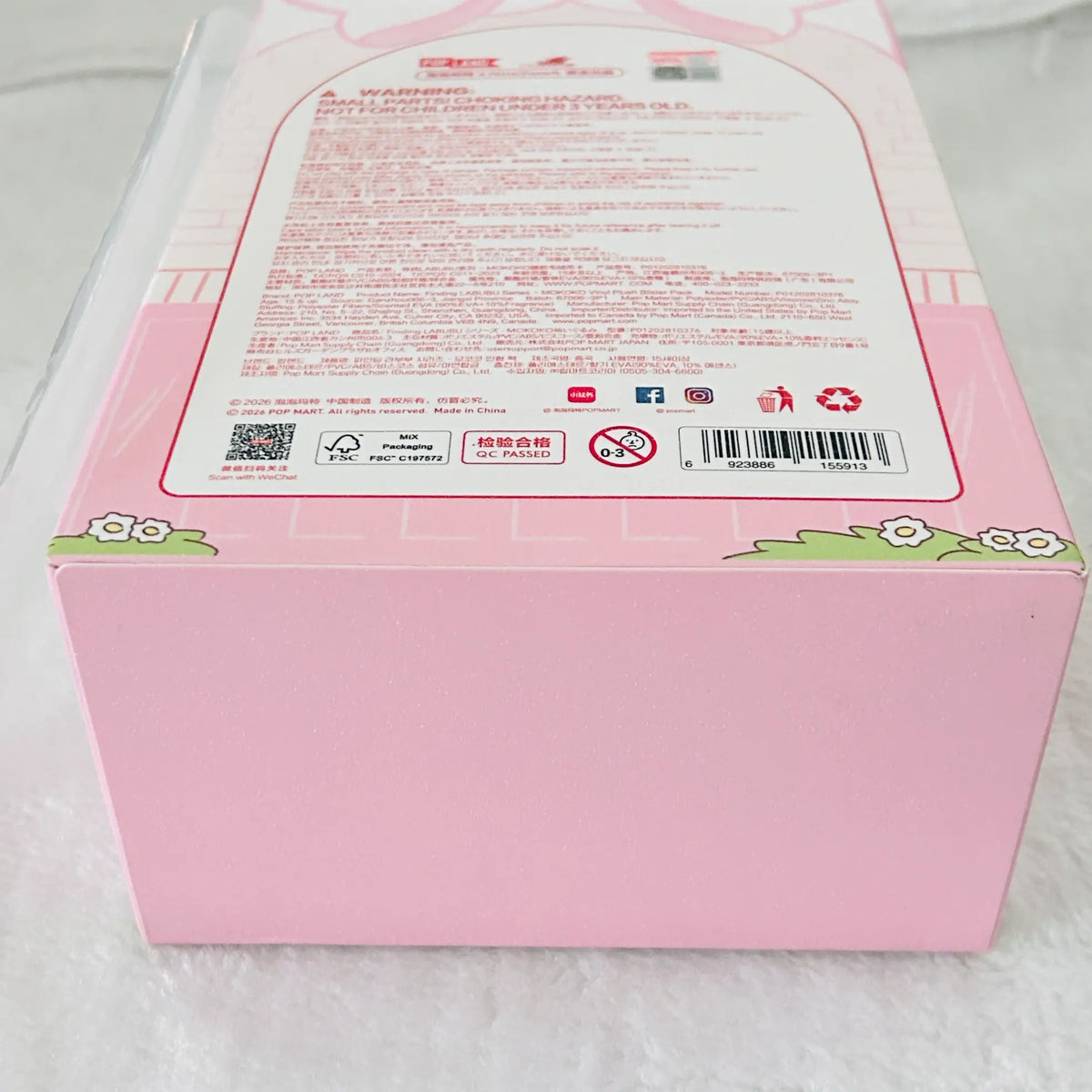 Pink product packaging box with a large white label containing red text, barcodes, and small cartoon floral details on the front and top, photographed on a white textured surface.