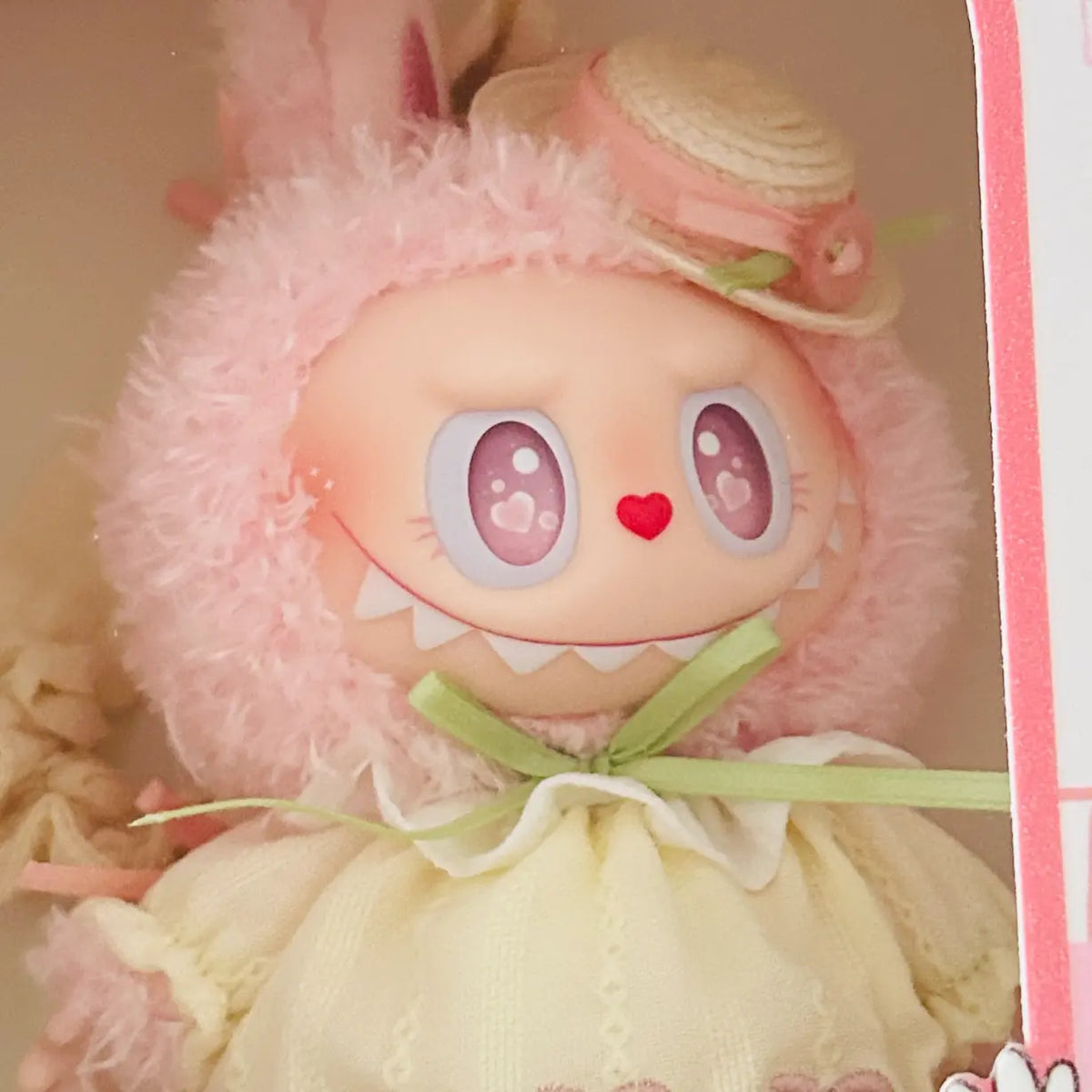 A close-up of a cute pink plush doll with large cartoon eyes, a red heart on its nose, and a pale yellow dress, shown inside a box with pink packaging visible on the right side.