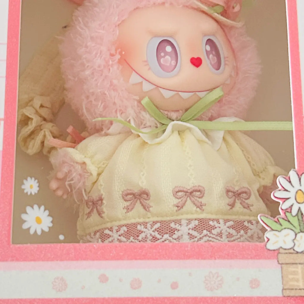 A cute pink plush doll with large cartoonish eyes, a red heart nose, and a fuzzy pink hood, dressed in a cream-colored embroidered dress with pink bows and lace trim, presented inside a decorative pink display box.