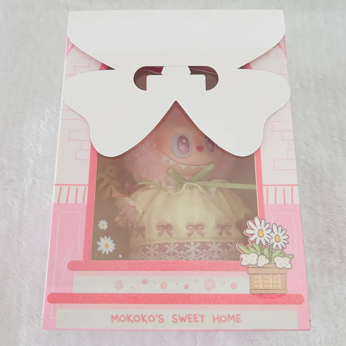 A packaged plush doll toy inside a pink decorative gift box with a die-cut bow window. The doll has a pink face and body, wearing a cream-colored dress with a green bow.