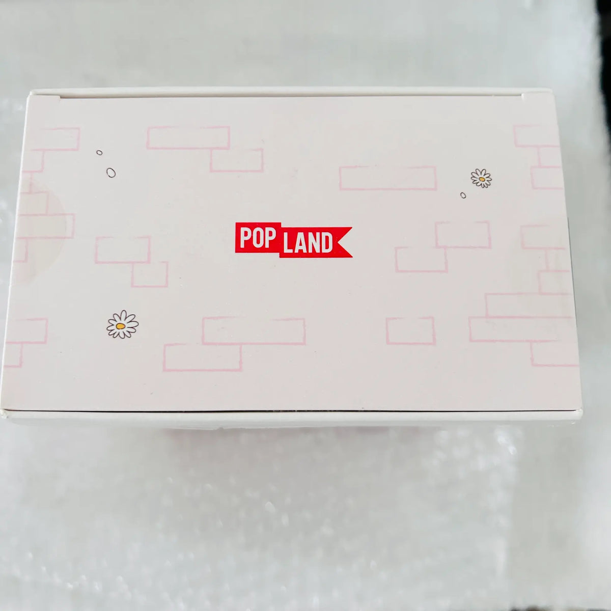 A square white product box with a red 'POP LAND' logo on the lid, decorated with light pink block patterns and small flower illustrations, resting on a white textured surface.
