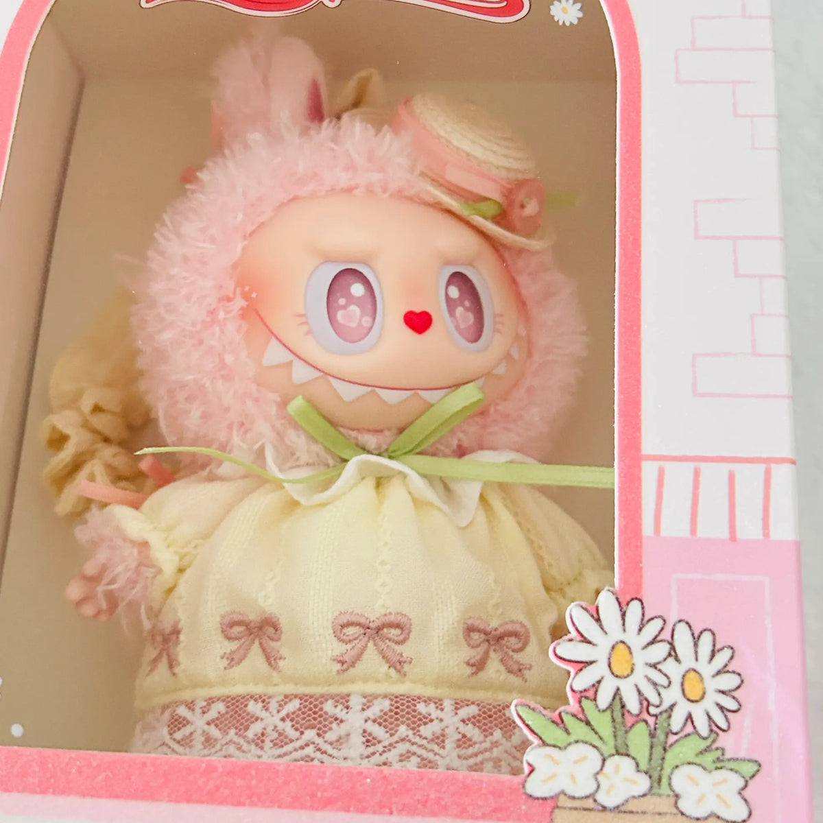 A cute plush doll with a fuzzy pink bunny hood and a cream-colored dress decorated with pink bows, shown inside its pink decorative retail box.