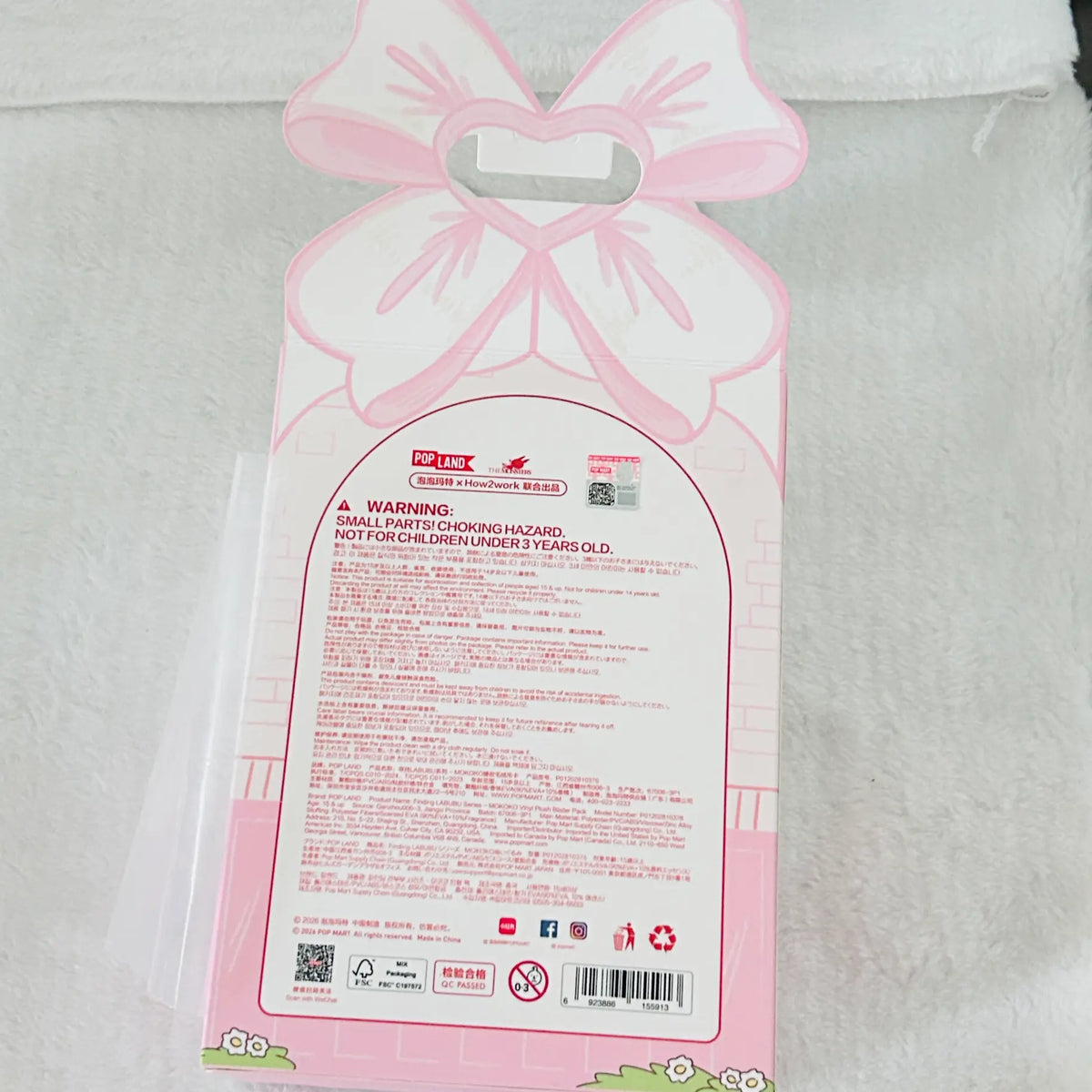 Pink and white decorative gift bag packaging with a bow-shaped handle and a large warning label on the front, photographed on a white textured fabric background.