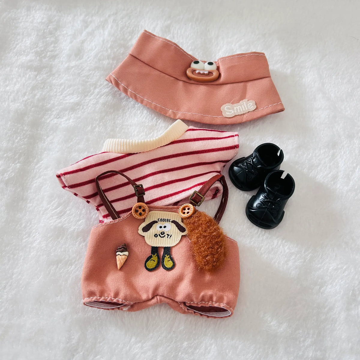 A doll outfit set laid flat on a white textured background, including a dusty coral overalls, a red-and-white striped top, and black boots. 