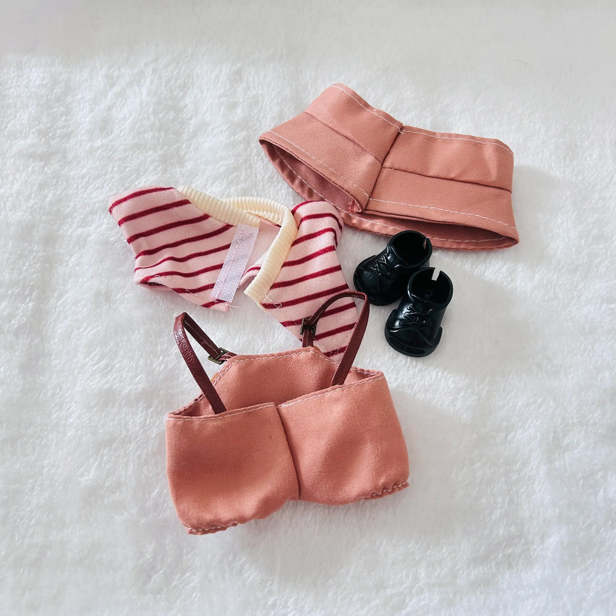 A doll outfit set laid flat on a white textured surface, including a peach satin-like top and skirt, a red-and-white striped bodysuit, a matching peach headband, and small black shoes.