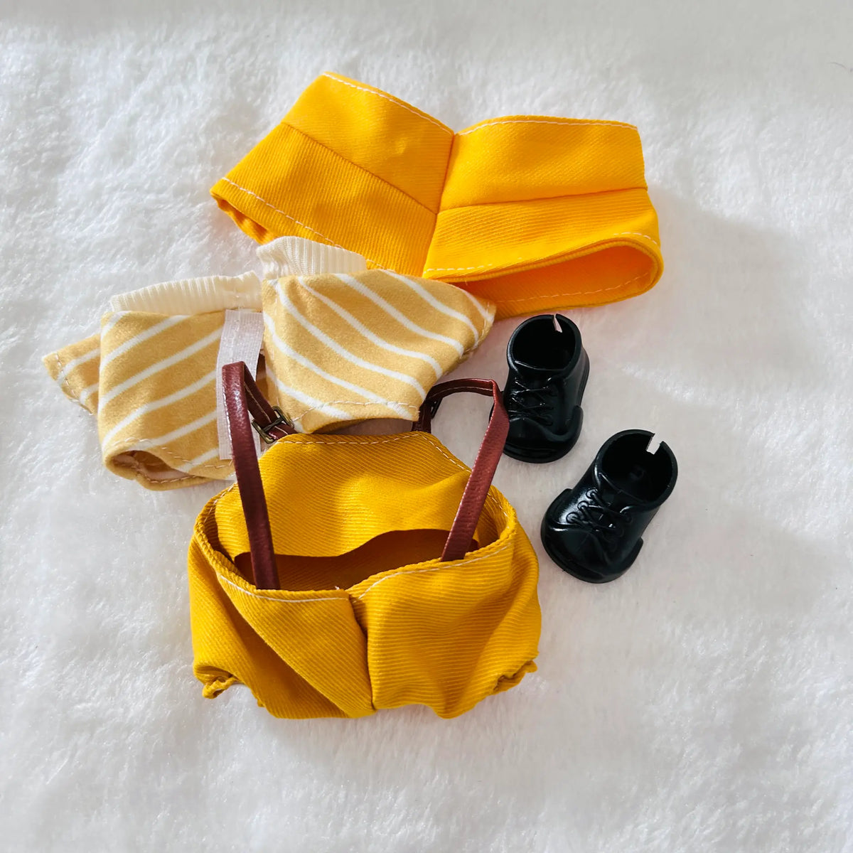 A doll outfit set laid flat on a white textured surface, including a yellow skirt, a yellow-and-white striped top, a yellow mini skirt with brown straps, and a pair of small black shoes.