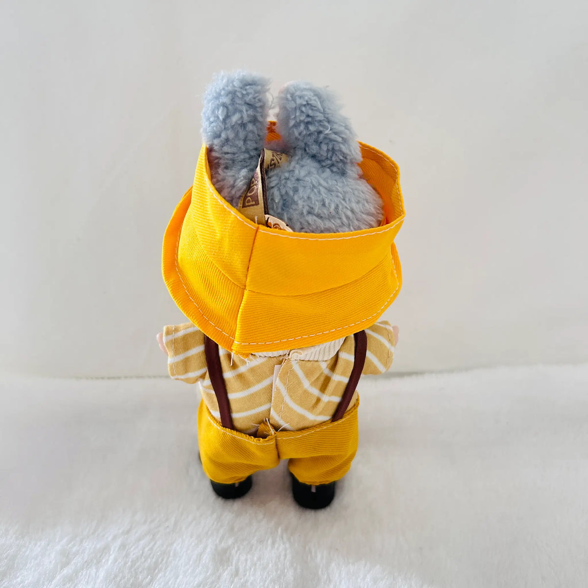 A back view of a Labubuwearing a bright yellow hat, yellow pants, brown suspenders, and a yellow-and-white striped shirt.