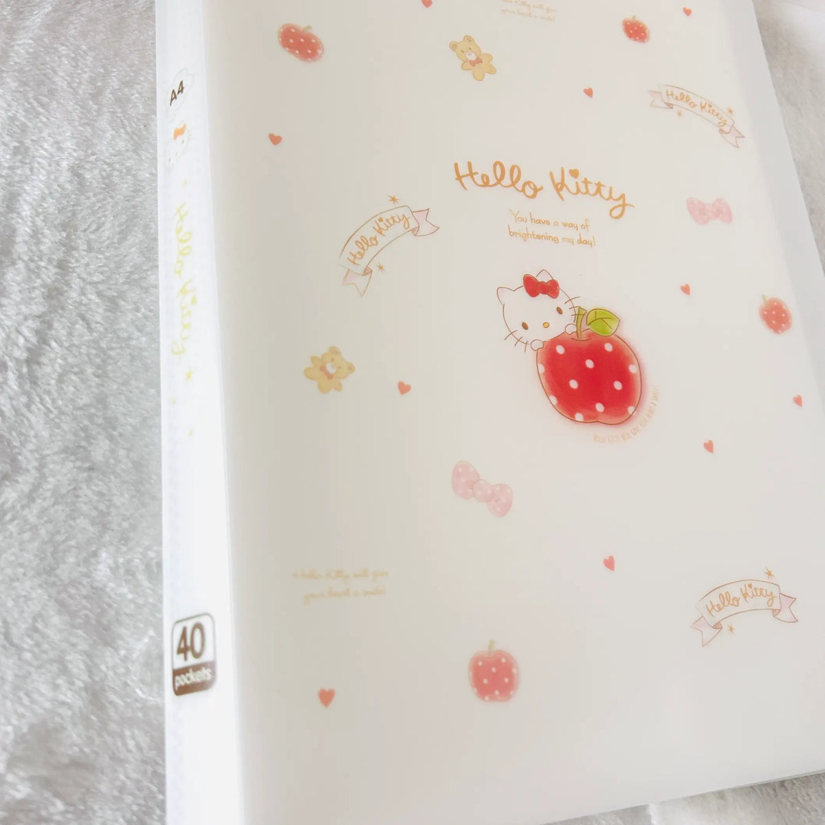 A translucent white plastic file folder featuring Hello Kitty designs, including a central apple illustration, 'Hello Kitty' text, small hearts, bows, and apples.