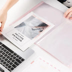 A translucent pink plastic photo organiser or binder sheet on a white desk, with hands holding it open and a printed card inserted.  