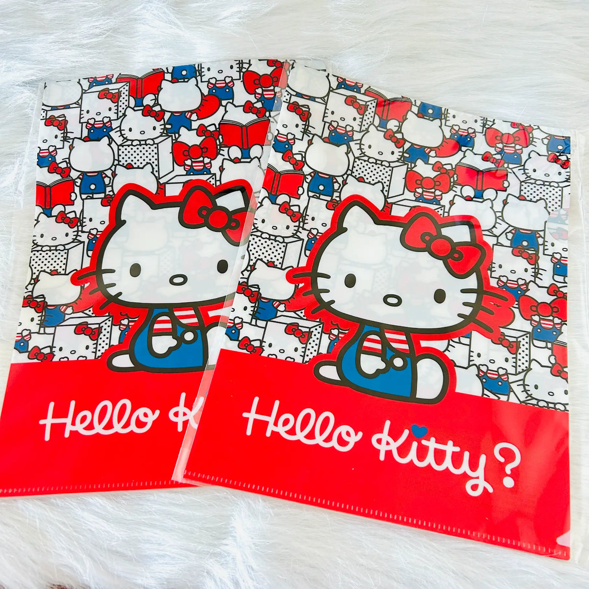 Two Hello Kitty file folders on a white faux fur surface. The folders feature a busy red, white, and blue patterned background with a large graphic of Hello Kitty in the center and a solid red bottom band with 'Hello Kitty?' written in white script.