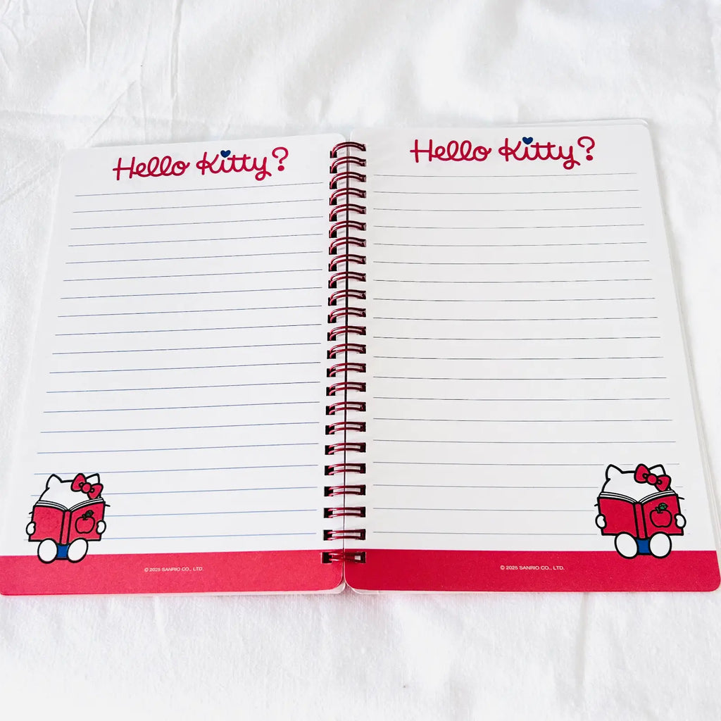 An open spiral-bound Hello Kitty writing notebook on a white fabric background. The pages are lined with 'Hello Kitty?' written in red at the top of each page, and cartoon illustrations of Hello Kitty appear in the bottom corners.