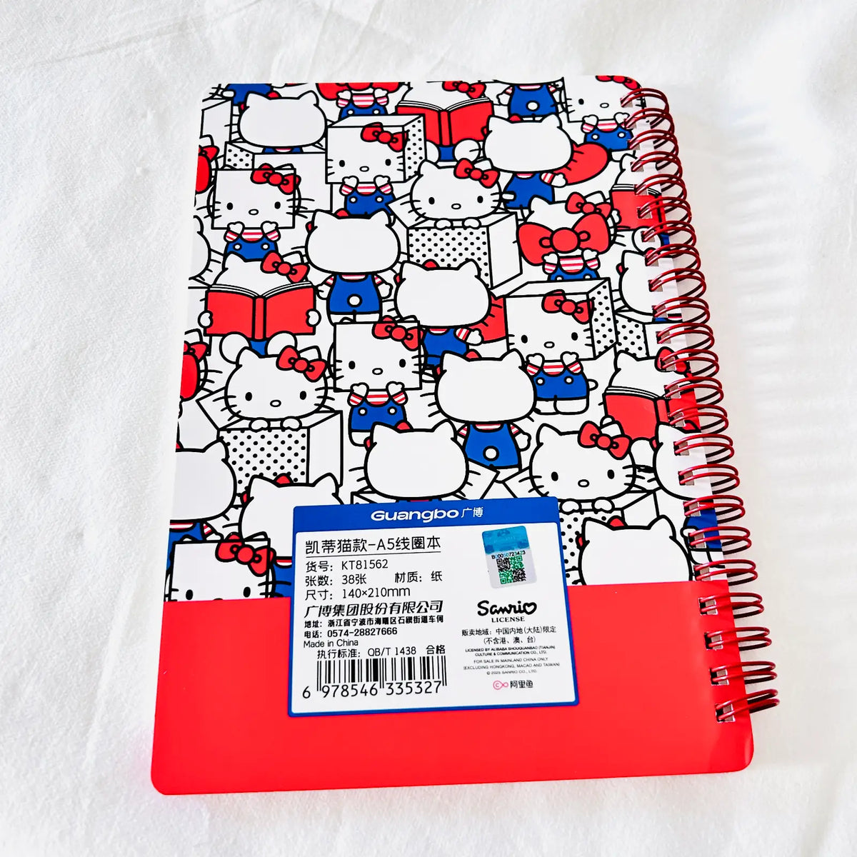 A spiral-bound notebook with a white cover covered in a repeating Hello Kitty pattern in red, blue, black, and white, featuring a solid red lower section and a blue product label on the front.