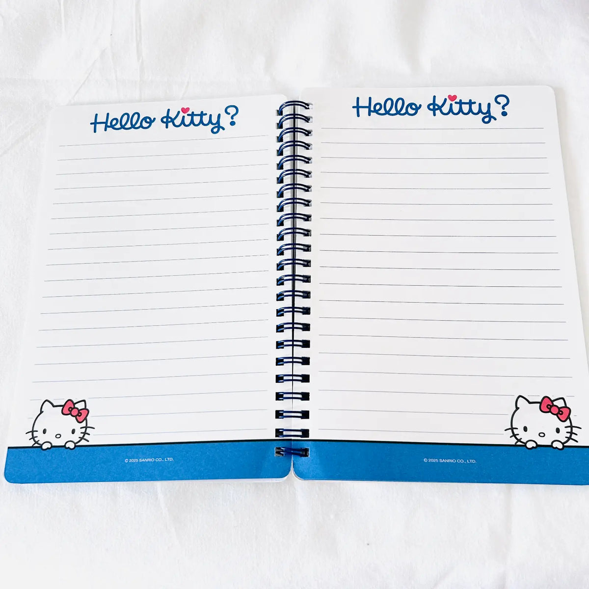 An open Hello Kitty spiral notebook with lined pages, blue 'Hello Kitty?' text at the top of each page, and Hello Kitty illustrations in the bottom corners, photographed on a white fabric background.