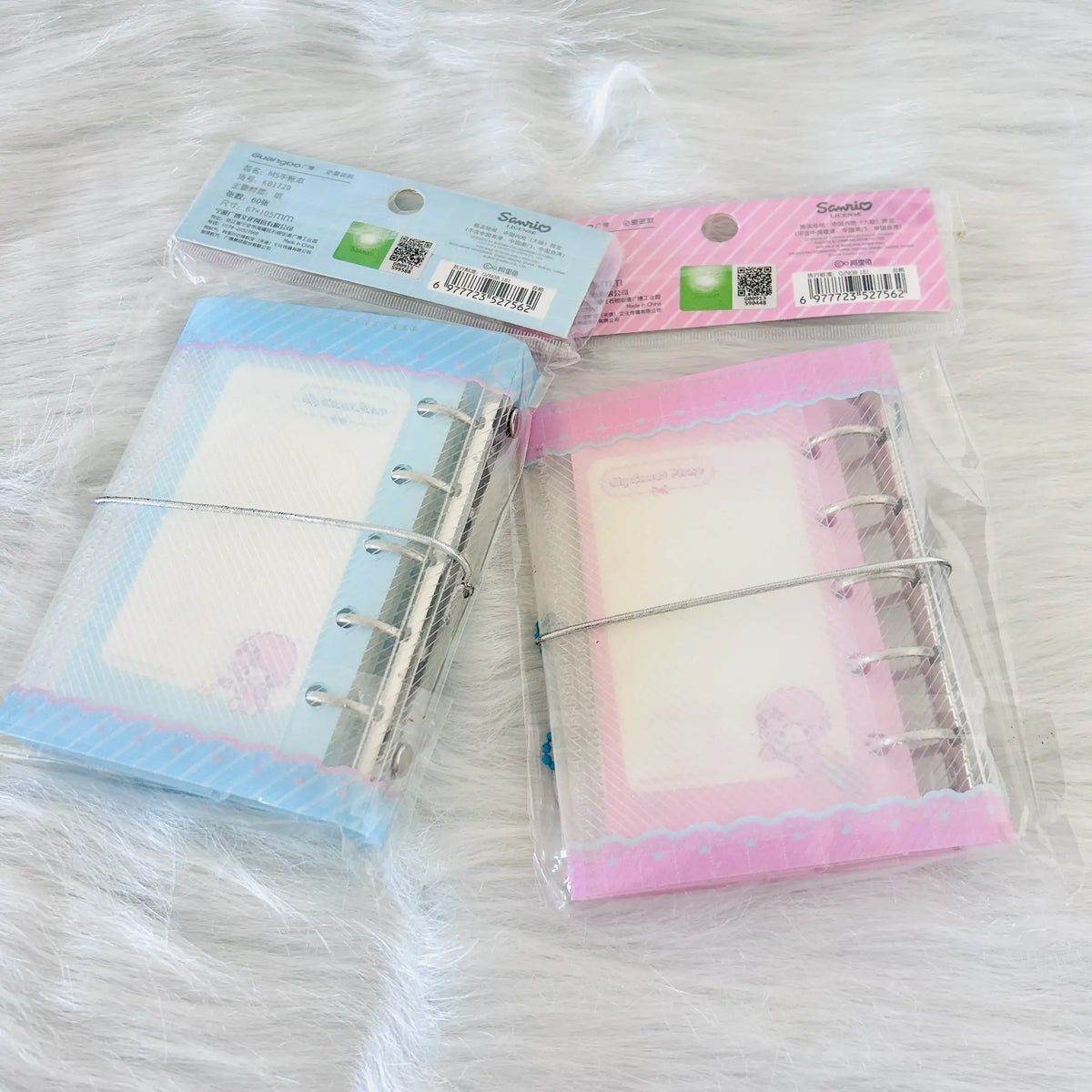Two small ring-bound planner inserts or notepad covers in clear plastic packaging, one light blue and one pink, placed on a white faux-fur surface. The packaging shows the Sanrio anti-counterfeiting label. 
