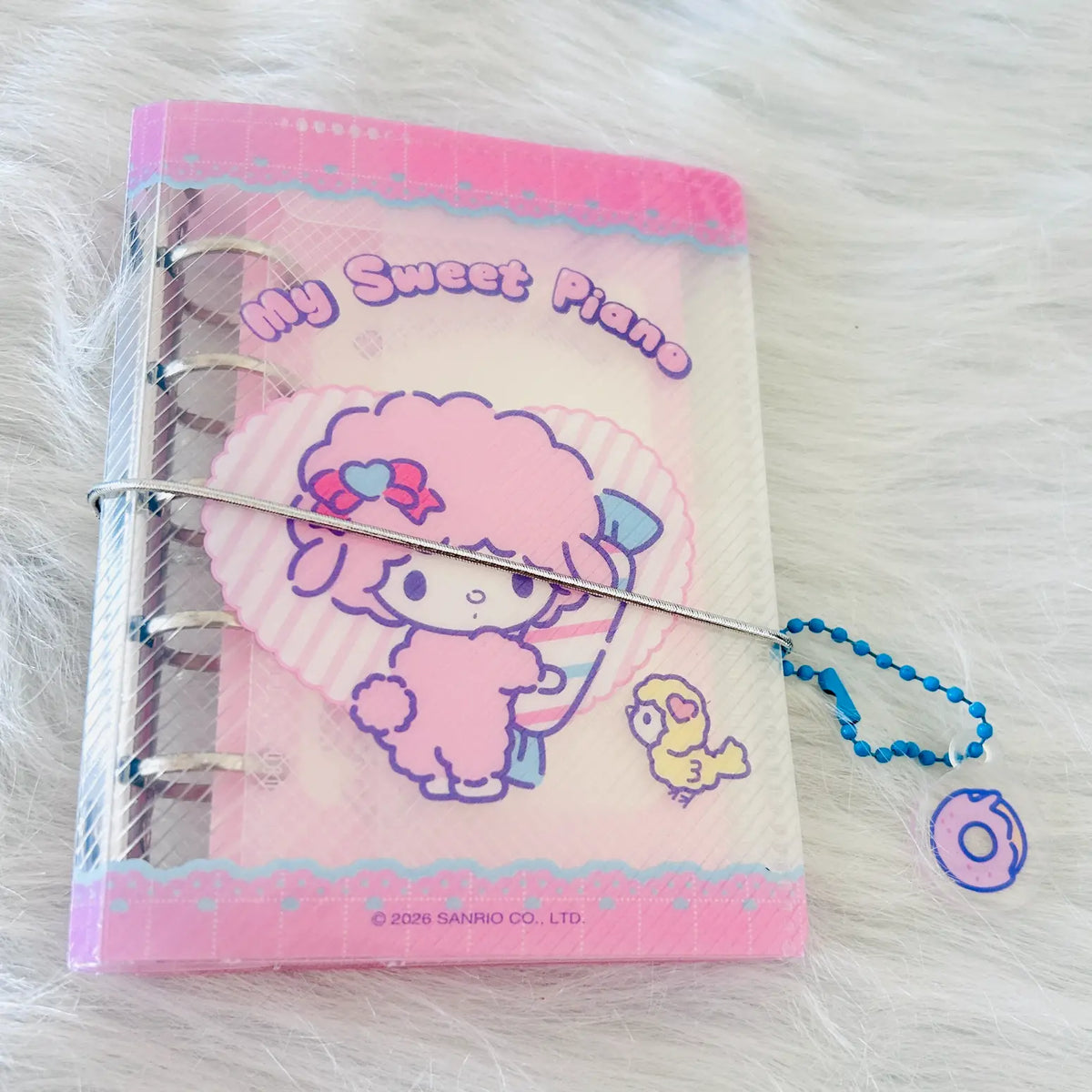 A Sanrio 'My Sweet Piano' ring-bound notebook featuring a pink sheep character and a small yellow bird, shown on a white faux fur background with a blue bead chain attached.