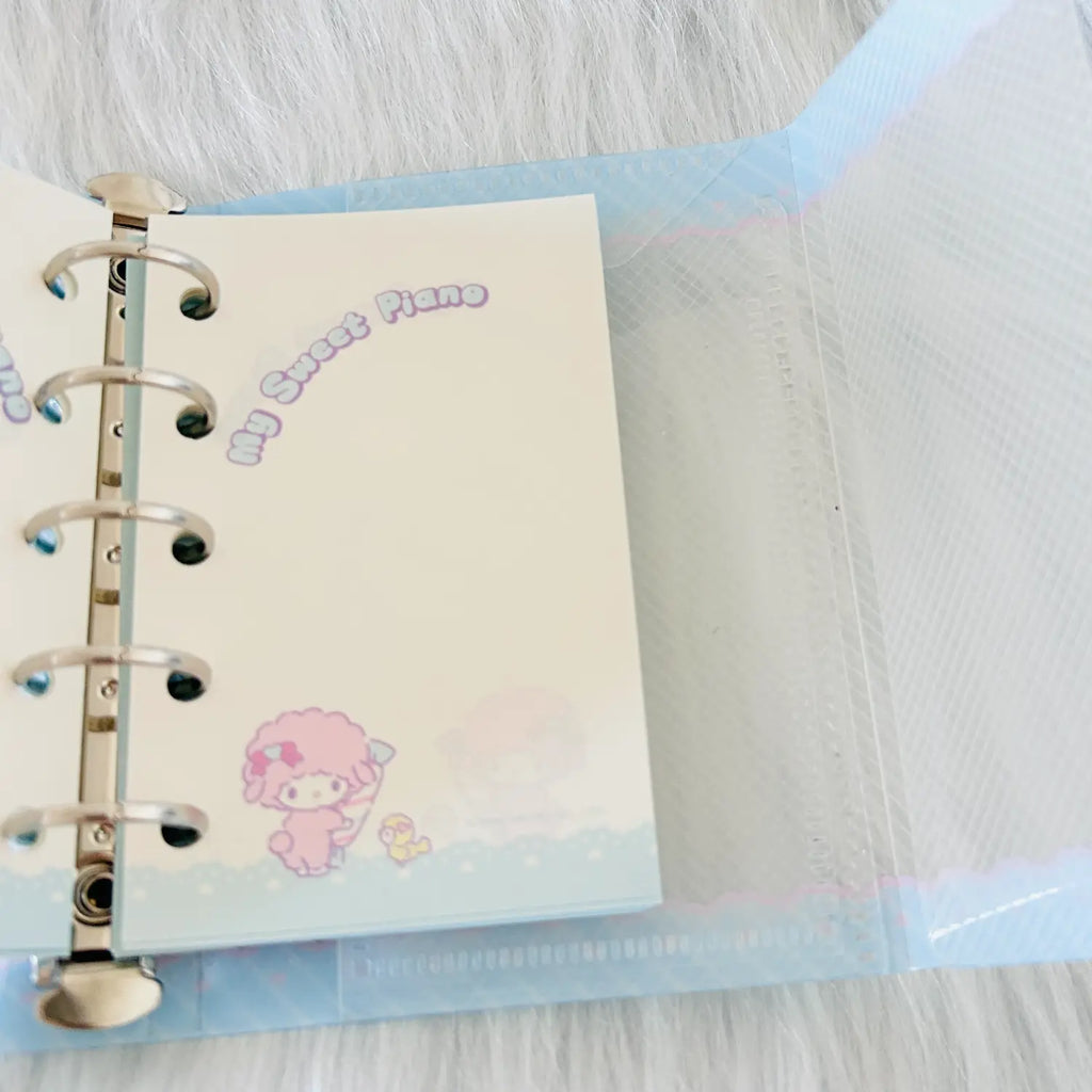 A close-up of a pastel-colored ring binder notebook with a clear plastic cover, showing a cream page with a pink cartoon character and the text 'My Sweet Piano'.