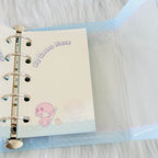 A close-up of a pastel-colored ring binder notebook with a clear plastic cover, showing a cream page with a pink cartoon character and the text 'My Sweet Piano'.