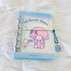A Sanrio 'My Sweet Plane' ring binder planner or notebook featuring a pink penguin character on a light blue and white cover, wrapped with a silver string and a blue bead chain, resting on a white faux fur background.
