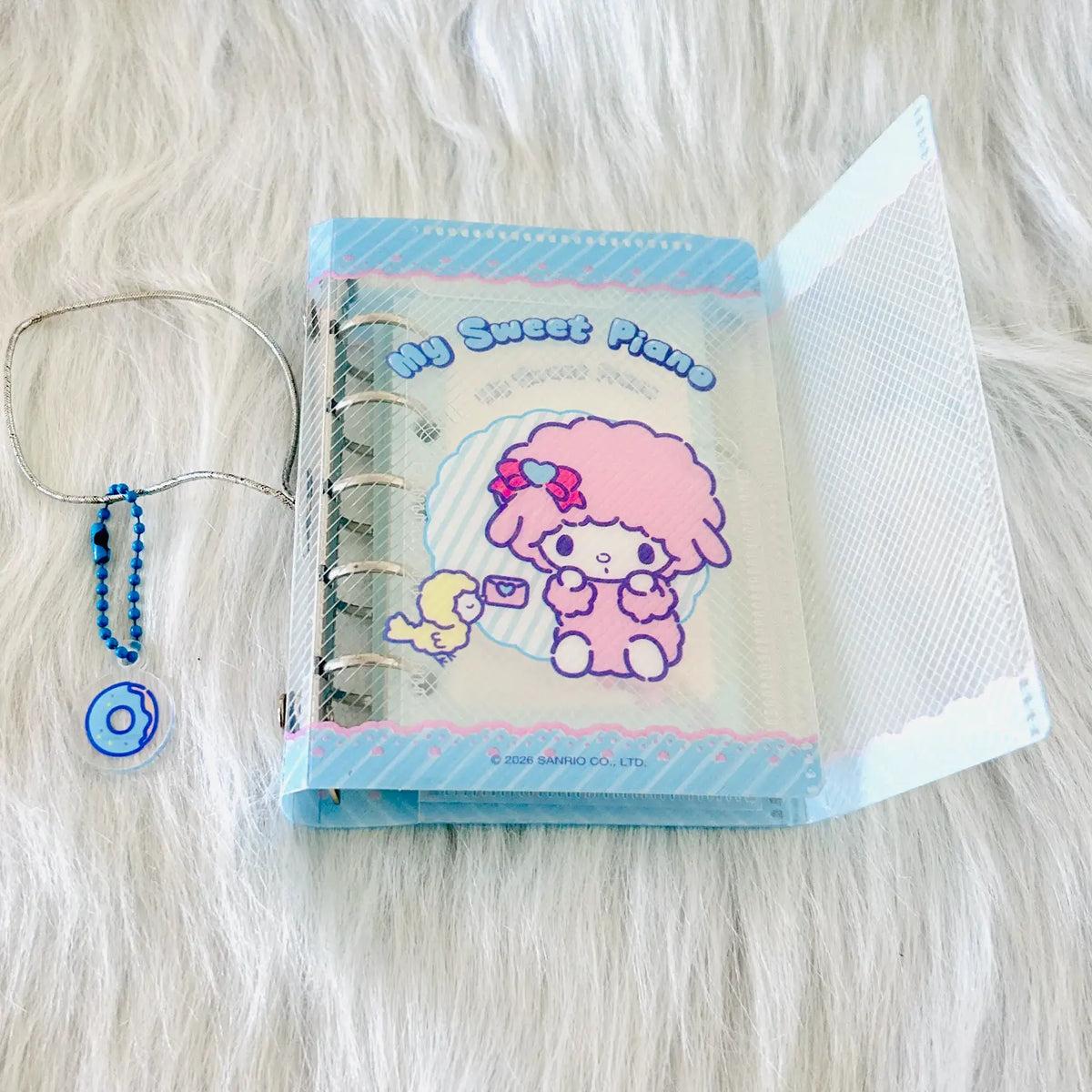 A Sanrio 'My Sweet Piano' ring binder notebook with a pink-haired character illustration, displayed on a white faux fur background alongside a blue beaded chain.